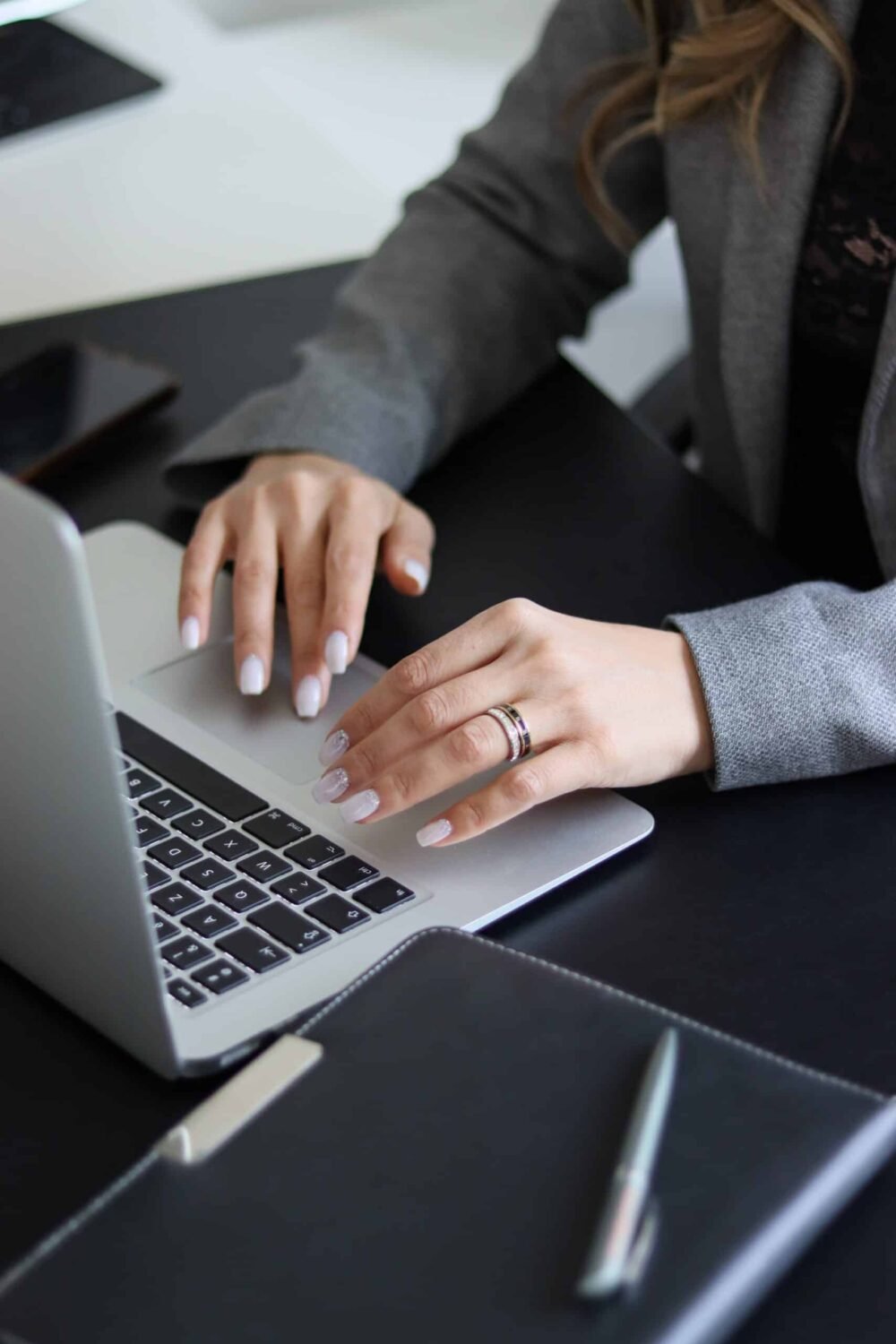 A person in a gray blazer typing on a laptop at a black desk, with a notebook and pen nearby, working to boost your website's SEO. Only their hands and arms are visible.