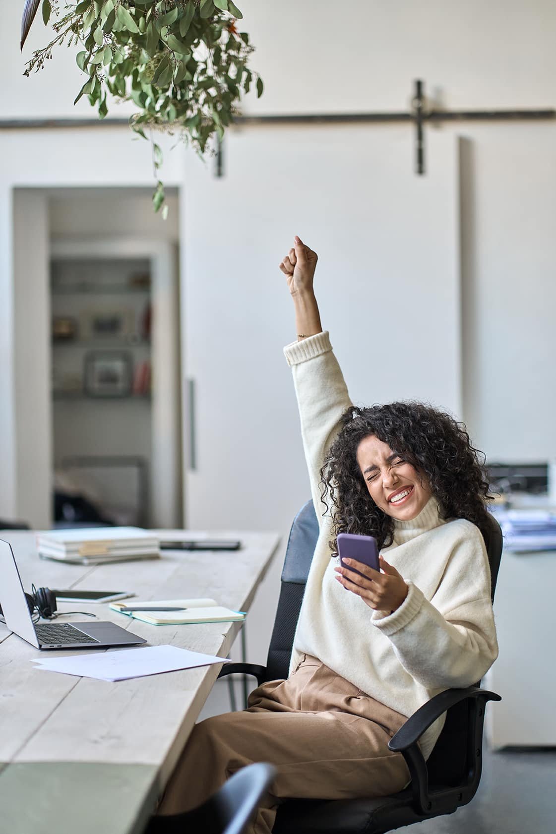 A joyful woman with curly hair stretches her arm up while holding a smartphone, sitting at an office desk surrounded by papers addressing top WordPress issues.