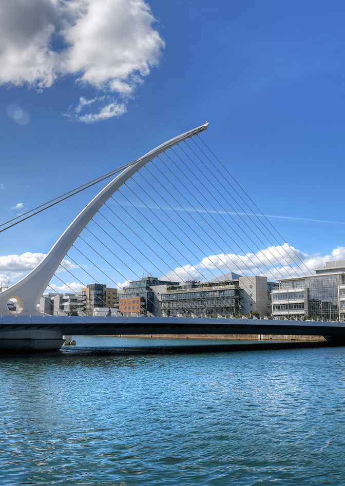 A modern white suspension bridge with cable supports spans over a body of water, with urban buildings in the background under a partly cloudy blue sky.